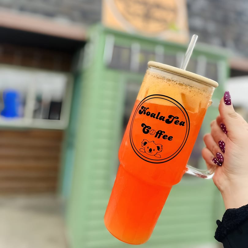 A hand holds a KoalaTea Coffee branded glass with a vibrant orange drink and foam.