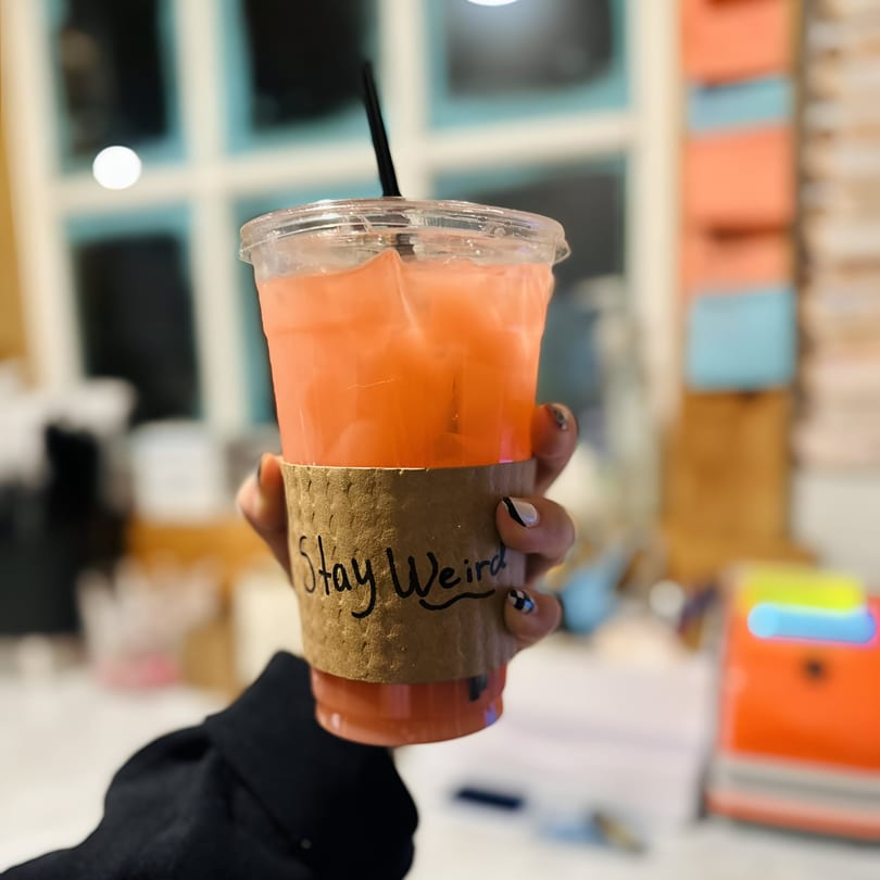 Hand holding a KoalaTea Coffee drink with a "Stay Weird" sleeve, against a blurred window background.